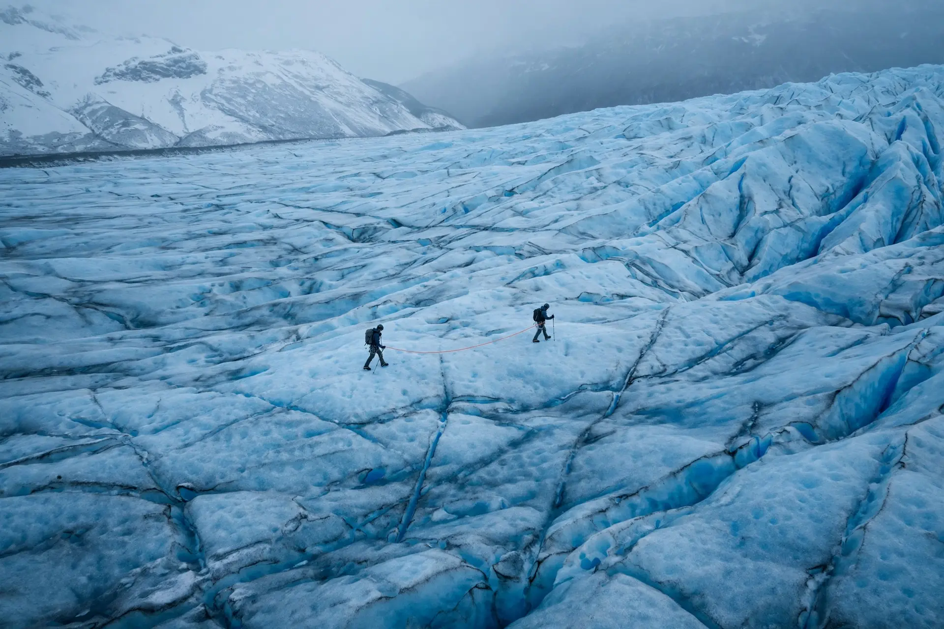 Two climbers crossing a vast blue glacier, small against the ice.